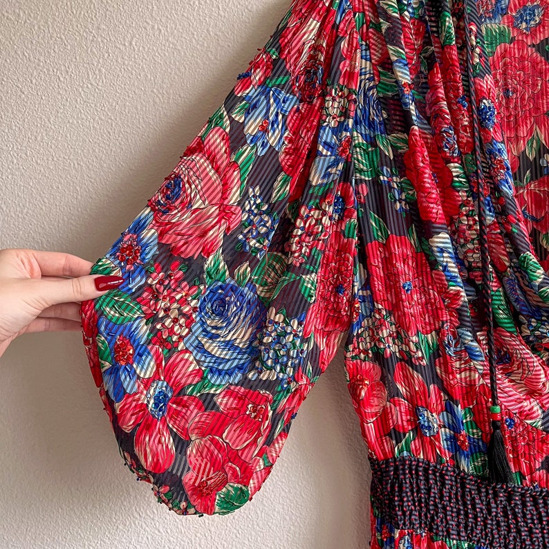 K&ouml;nnte beinhalten: Ein schwarzes und rotes Blumenkleid mit R&uuml;schen am Ausschnitt und kurzen &Auml;rmeln. Das Kleid hat ein tailliertes Oberteil und einen flie&szlig;enden Rock.