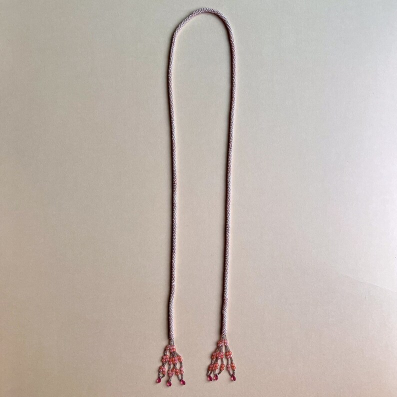 May include: A long, thin, beaded necklace with pink and white beads. The necklace has two tassels at the ends, each with pink and white beads.
