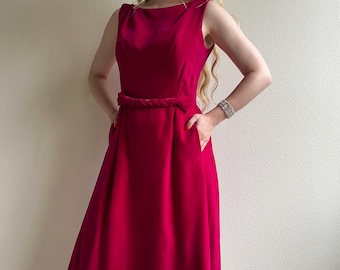 May include: A sleeveless, knee-length dress in a vibrant magenta hue. The dress features a boat neckline, a braided belt, and pockets. The model is wearing a silver bracelet. The dress is a classic, elegant style.