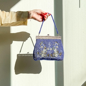 K&ouml;nnte beinhalten: Eine blaue Perlenhandtasche mit einem silbernen Verschluss und Blumenstickerei. Die Tasche hat einen kurzen Henkel und wird von einer Hand mit rotem Nagellack gehalten.