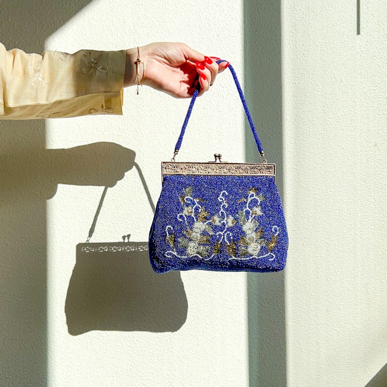 K&ouml;nnte beinhalten: Eine blaue Perlenhandtasche mit einem silbernen Verschluss und Blumenstickerei. Die Tasche hat einen kurzen Henkel und wird von einer Hand mit rotem Nagellack gehalten.