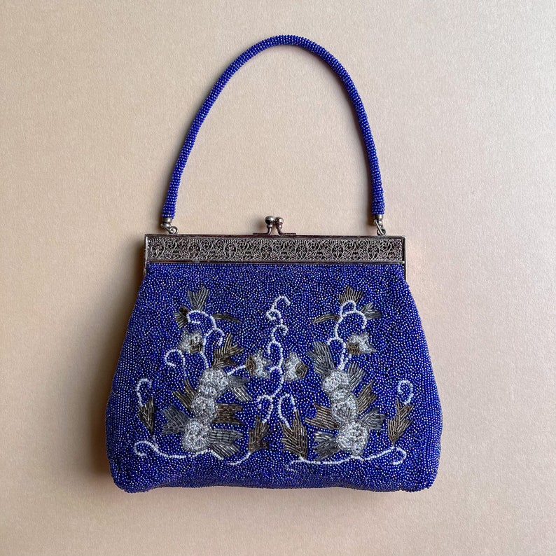 K&ouml;nnte beinhalten: Eine blaue Perlen-Abendtasche mit silbernem Metallrahmen und Blumenstickerei. Die Tasche hat einen oberen Henkel.