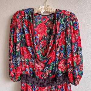 K&ouml;nnte beinhalten: Ein rotes, blaues und gr&uuml;nes Blumenkleid mit gerafftem Ausschnitt und einem schwarzen elastischen Bund. Das Kleid hat lange &Auml;rmel mit einem gerafften Puff an der Schulter.