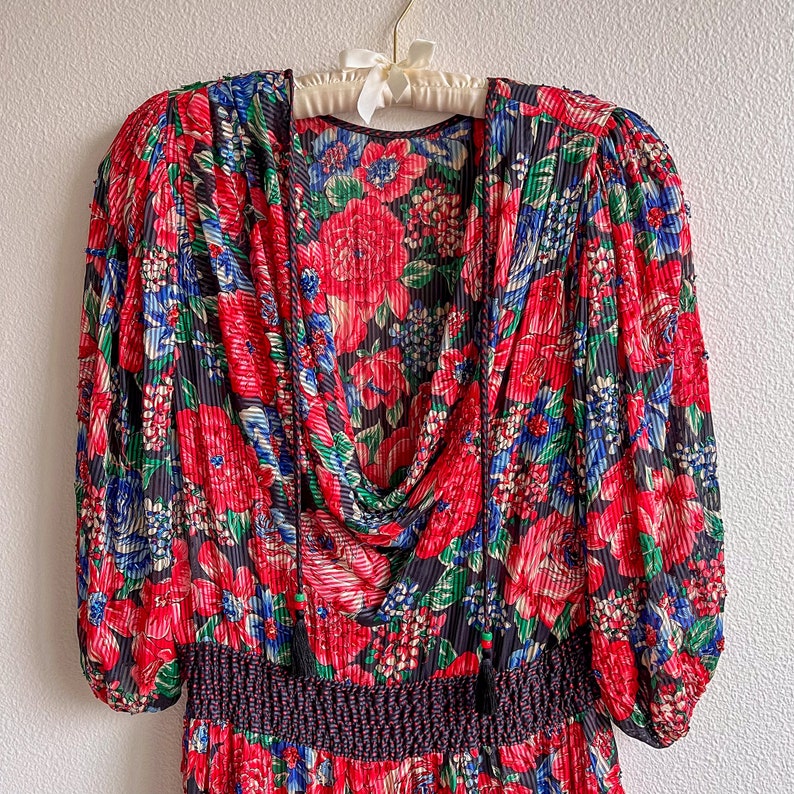 K&ouml;nnte beinhalten: Ein rotes, blaues und gr&uuml;nes Blumenkleid mit gerafftem Ausschnitt und einem schwarzen elastischen Bund. Das Kleid hat lange &Auml;rmel mit einem gerafften Puff an der Schulter.