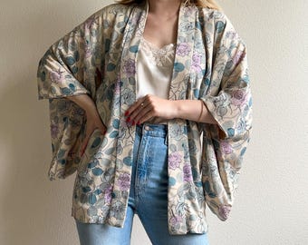 Könnte beinhalten: Ein beiger Kimono mit einem Blumenmuster aus blauen und lila Rosen. Der Kimono hat weite Ärmel und ist vorne offen. Das Model trägt ein weißes Camisole und blaue Jeans.