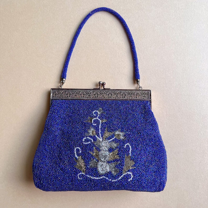 K&ouml;nnte beinhalten: Eine blaue Perlen-Abendtasche mit einem silbernen Verschluss und floraler Stickerei. Die Tasche hat einen oberen Henkel und einen Verschluss.