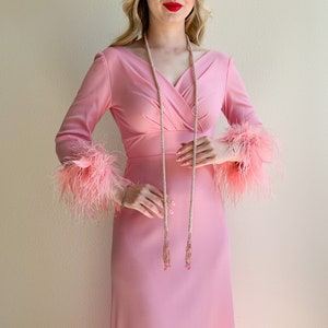 May include: A pink dress with long sleeves and feather trim. The dress has a V-neckline and a wrap-style bodice. The model is wearing a long, beaded necklace with pink accents.