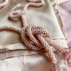 May include: A delicate pink pearl necklace with a knotted design, draped on a soft, light pink fabric background.