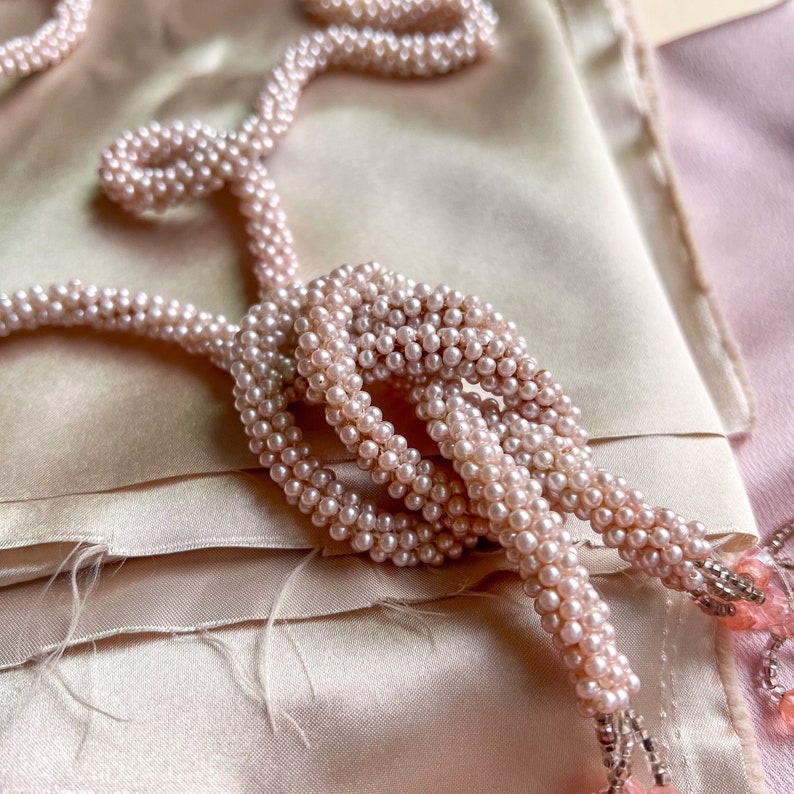 May include: A delicate pink pearl necklace with a knotted design, draped on a soft, light pink fabric background.