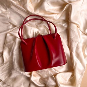 Könnte beinhalten: Eine leuchtend rote Handtasche mit zwei geschwungenen Henkeln. Die Tasche hat eine rechteckige Form mit einem gerafften Design auf der Vorderseite. Die Tasche ist aus glänzendem Material gefertigt und liegt auf einem cremefarbenen, seidigen Stoff.