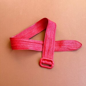May include: A bright pink woven belt with a rectangular buckle. The belt is made of a textured material and is displayed against a brown background. The belt's design is simple and elegant.