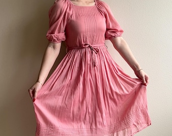 Könnte beinhalten: Ein rosa Baumwollkleid mit einem gerafften Rock und einer taillierten Taille. Das Kleid hat einen quadratischen Ausschnitt und kurze Puffärmel.