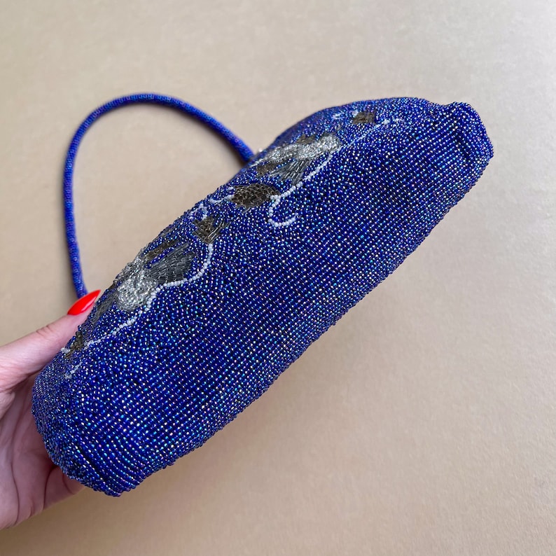 K&ouml;nnte beinhalten: Eine kleine blaue Perlenhandtasche mit silbernem Blumendesign. Die Tasche hat einen einzigen Riemen.