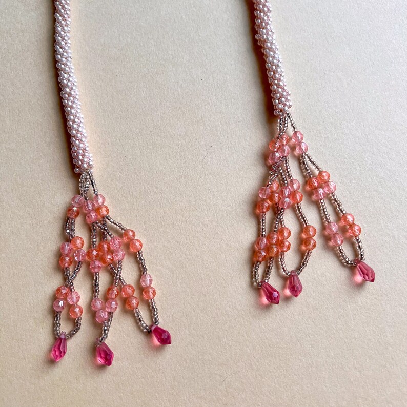 May include: Two strands of pink and peach beaded necklaces with pink and peach tassel pendants.