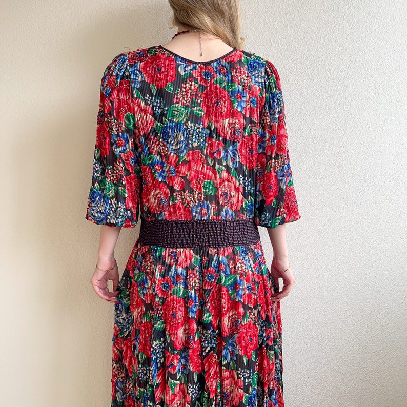 K&ouml;nnte beinhalten: Ein langes, rot, blau und gr&uuml;n gemustertes Blumenkleid mit einem schwarzen elastischen Bund. Das Kleid hat 3/4 &Auml;rmel und eine lockere, flie&szlig;ende Silhouette.