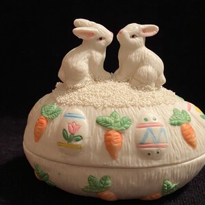 May include: White ceramic Easter egg-shaped trinket box with two white bunny figurines on top. The box is decorated with colorful Easter eggs, carrots, and green leaves.