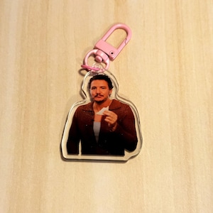 May include: A clear acrylic keychain featuring a color image of a person holding a small white object. The keychain has a pink clasp. The person is wearing a brown jacket and a light blue shirt.