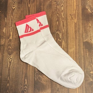 以下が含まれることがあります： ピンクのストライプとピンクの三角形のロゴが付いた白いアンクルソックスのペアです。