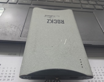 以下が含まれることがあります： 表面がテクスチャ加工された長方形のグレーのポータブルパワーバンク。デバイスの上部には「ROCKZ」および「By Zikcord」の文字が印刷されています。前面には2つのUSBポートと小さなインジケーターライトがあり、側面には4つの縦のドットが配置されています。