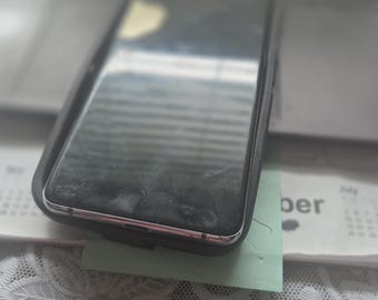 May include: A rectangular smartphone with a black screen and a dark protective case. The device is positioned at an angle, showing a metallic silver frame along the side edges. The screen surface appears reflective and shows faint smudges.