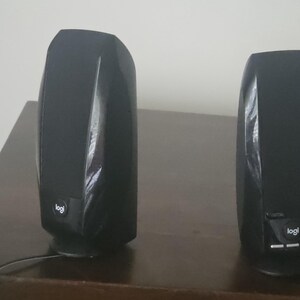 May include: A pair of black computer speakers with a glossy finish and a fabric mesh front. Each speaker features a small circular logo with the text logi at the bottom center. The right speaker includes three horizontal control buttons below the logo.