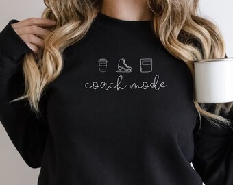 May include: Black sweatshirt with white text that reads "coach mode" and three icons: a coffee cup, ice skates, and a notebook. The model is wearing the sweatshirt and holding a white mug.