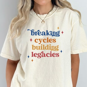 May include: A cream-colored t-shirt with the text "breaking cycles building legacies" in blue, orange, and red lettering with small star accents.