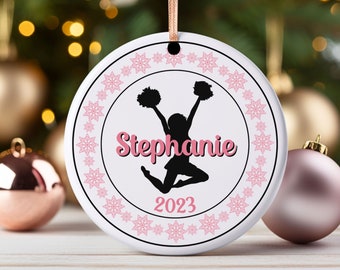 May include: White ceramic Christmas ornament with a pink snowflake border. The center features a black silhouette of a cheerleader with pom-poms, the name "Stephanie" in pink script, and the year "2023".