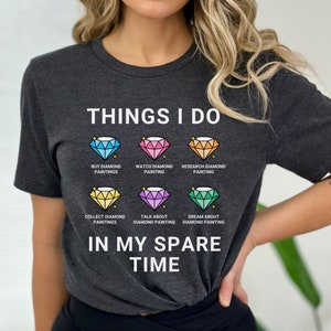 May include: A dark gray t-shirt with the text "THINGS I DO" and a series of colorful diamond illustrations. Each diamond has a different activity related to diamond painting, such as "BUY DIAMOND PAINTINGS", "WATCH DIAMOND PAINTING", "RESEARCH DIAMOND PAINTING", "COLLECT DIAMOND PAINTINGS", "TALK ABOUT DIAMOND PAINTING", and "DREAM ABOUT DIAMOND PAINTING". The text "IN MY SPARE TIME" is printed below the diamonds.
