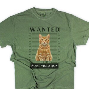 May include: A green t-shirt with a wanted poster design featuring a cartoon orange tabby cat. The poster says "WANTED" and "NOISE VIOLATION".