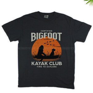 Könnte beinhalten: Schwarzes T-Shirt mit einer Grafik im Vintage-Stil von einem Bigfoot und einem Hund in einem Kajak. Die Grafik enthält den Text "Certified Bigfoot Kayak Club Time to Explore".