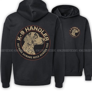 May include: A black hooded sweatshirt featuring a graphic on the back and front. The back displays a large circular design with a dog illustration, the text K-9 HANDLER, and the phrase BECAUSE HUMANS NEED HEROES TOO. The front has a smaller version.