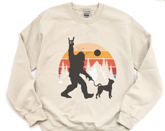 May include: A beige sweatshirt with a retro-style graphic of a Bigfoot with a dog on a leash. The graphic features a sunset, mountains, and trees. The Bigfoot is making a rock and roll hand gesture.