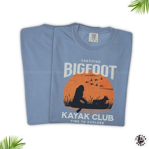 May include: A light blue t-shirt with a graphic design of a Bigfoot in a kayak with a dog. The text on the shirt reads "Certified Bigfoot Kayak Club Time to Explore".