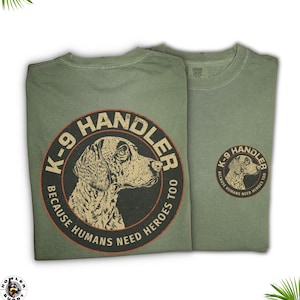 May include: Two olive green t-shirts. Each shirt displays a circular design with a dog illustration and the text "K-9 Handler Because Humans Need Heroes Too." One shirt has a smaller version of the design on the upper back.