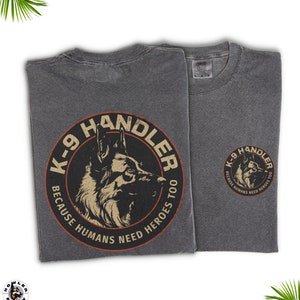 May include: A gray t-shirt with a black and orange circle graphic on the back. The graphic features a German Shepherd dog head and the text "K-9 Handler Because Humans Need Heroes Too".