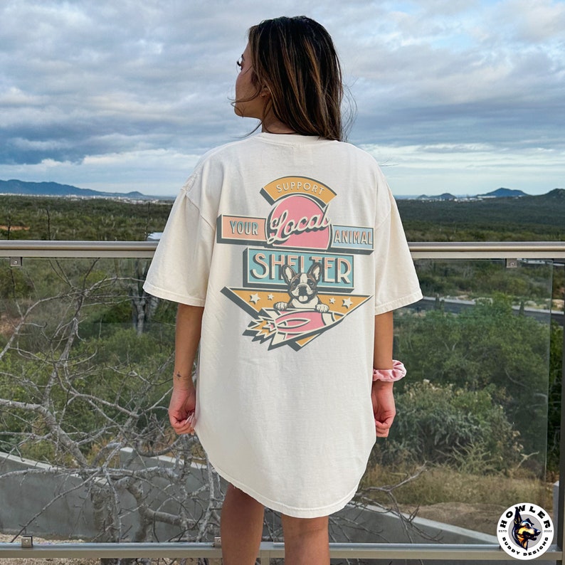 May include: Cream-colored t-shirt with a graphic that reads "Support Your Local Animal Shelter." The design features a dog on a surfboard with a retro color scheme. The shirt is worn by a person.