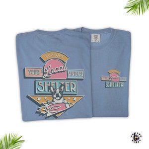May include: Two light blue t-shirts with a graphic supporting local animal shelters. The design features a French bulldog on a rocket, with text reading "Support Your Local Animal Shelter". One shirt is folded, the other is unfolded.