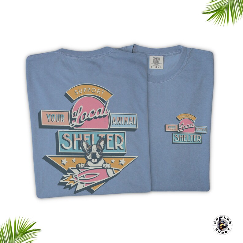May include: Two light blue t-shirts with a graphic supporting local animal shelters. The design features a French bulldog on a rocket, with text reading "Support Your Local Animal Shelter". One shirt is folded, the other is unfolded.