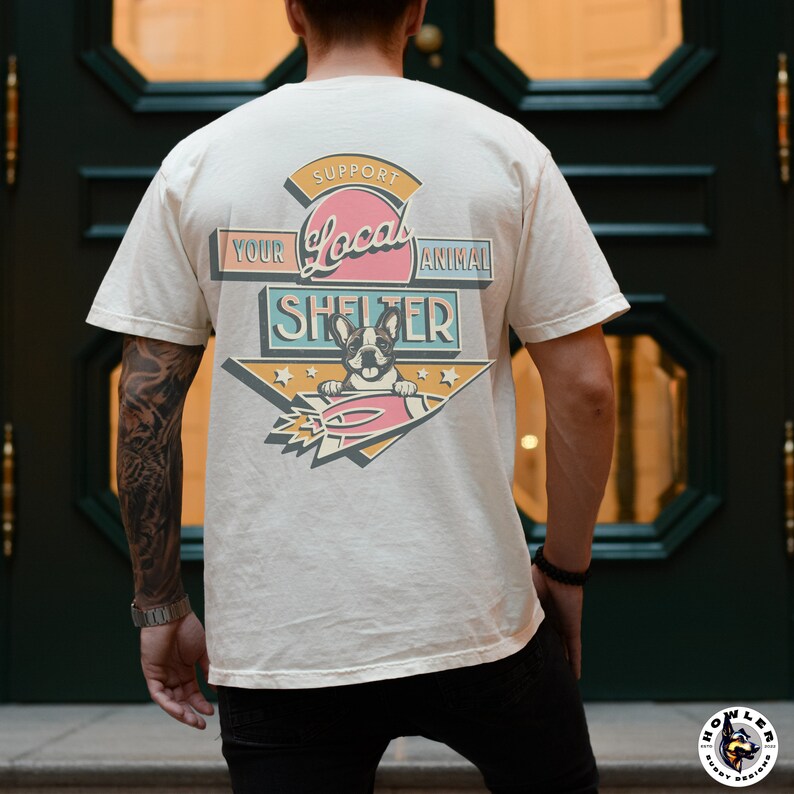May include: Cream-colored t-shirt with a graphic supporting local animal shelters. The design features a French bulldog, a rocket, and the text "Support Your Local Animal Shelter" in a retro style. The shirt is worn by a person.
