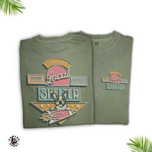 May include: Two olive green t-shirts with a graphic that reads "Support Your Local Animal Shelter." The design features a French bulldog in a rocket ship. The shirts are made of a soft material.