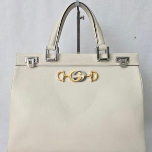 May include: A cream-colored leather handbag with a textured surface, featuring two top handles and silver-tone metal hardware. The front is adorned with a central gold and silver-tone metal horsebit-inspired emblem and two silver-tone side clasps.