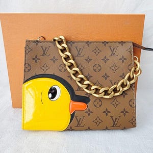 Könnte beinhalten: Braune und beige Handtasche mit goldenem Kettenriemen. Die Tasche hat ein Cartoon-Enten-Design in Gelb, Schwarz und Orange. Die Tasche hat ein sich wiederholendes Muster aus braunen Logos.