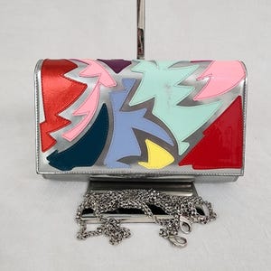 May include: A rectangular silver clutch bag featuring a metallic finish and an abstract pattern of appliqued shapes in red, pink, purple, light blue, dark blue, mint green, and yellow. The bag includes a detachable silver-tone metal chain strap.