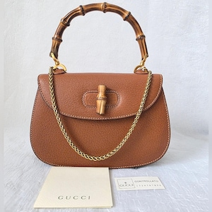 May include: A brown leather handbag featuring a curved bamboo top handle and a bamboo turn-lock closure. The bag includes a gold-tone chain shoulder strap and is accompanied by a Gucci brand card and a Controllato card with the text 1234567890.