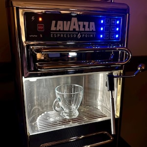 Könnte beinhalten: Eine silberne und schwarze Espressomaschine mit einer Glas Tasse auf dem Tropfblech. Die Maschine hat ein blaues digitales Display mit dem Text "Lavazza Espresso Point" auf der Vorderseite.