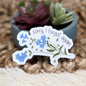 May include: A white sticker with blue forget-me-not flowers and the text "sorry I forgot again".