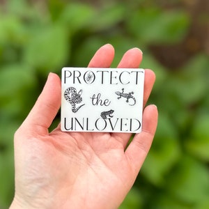May include: White sticker with black text and illustrations of a spider, snake, frog, and lizard. The text reads "Protect the Unloved".