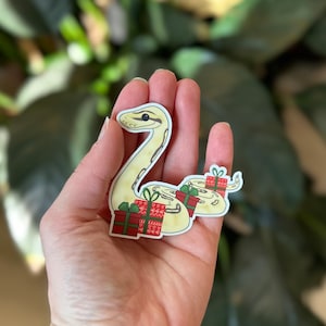 May include: A white and yellow snake with a red and white patterned gift box on its back. The snake is curled up in a festive pose.