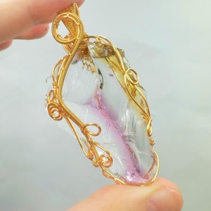 Puede incluir: Un colgante de cristal transparente facetado con un tono rosa y amarillo, envuelto en alambre dorado. El alambre forma un diseño delicado e intrincado alrededor del cristal.